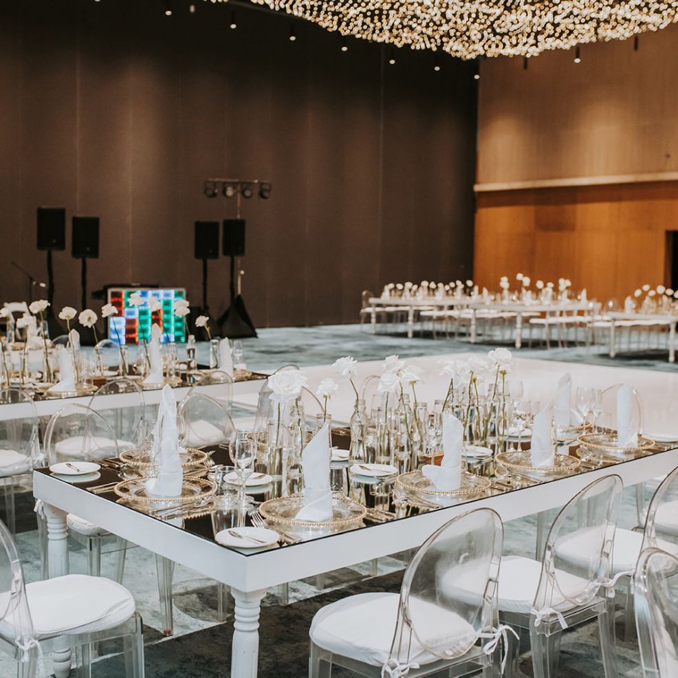 Banquet tables with white wedding decor and clear chairs set in a spacious destination wedding hall.