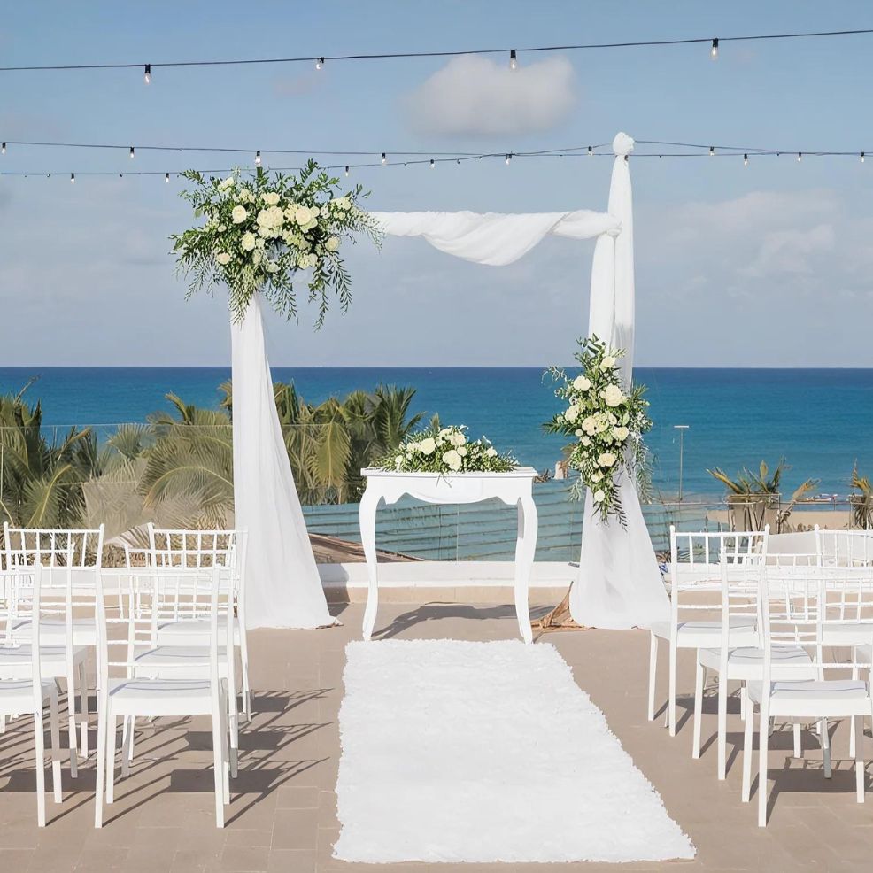Outdoor wedding setup with white chairs, a white aisle runner, and a floral arch decorated with white fabric and flowers, overlooking the ocean with a clear blue sky.