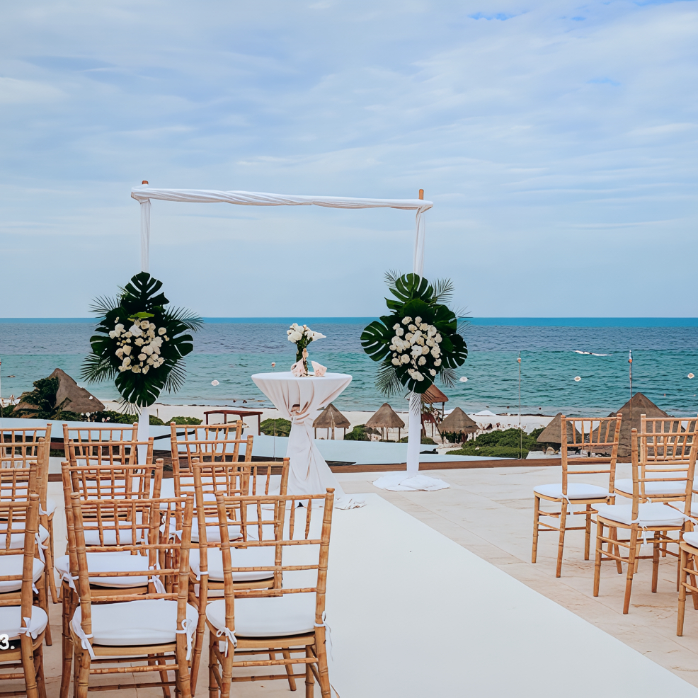 Destination wedding setup on the beach with wooden chairs and a floral arch overlooking the ocean.