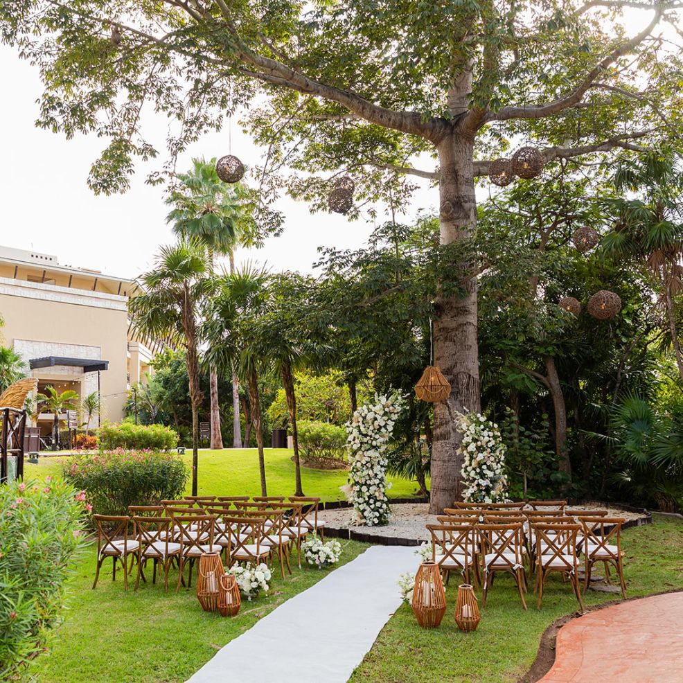 Destination wedding setup outdoors with rows of chairs, white aisle runner, and floral arch beneath a tree.
