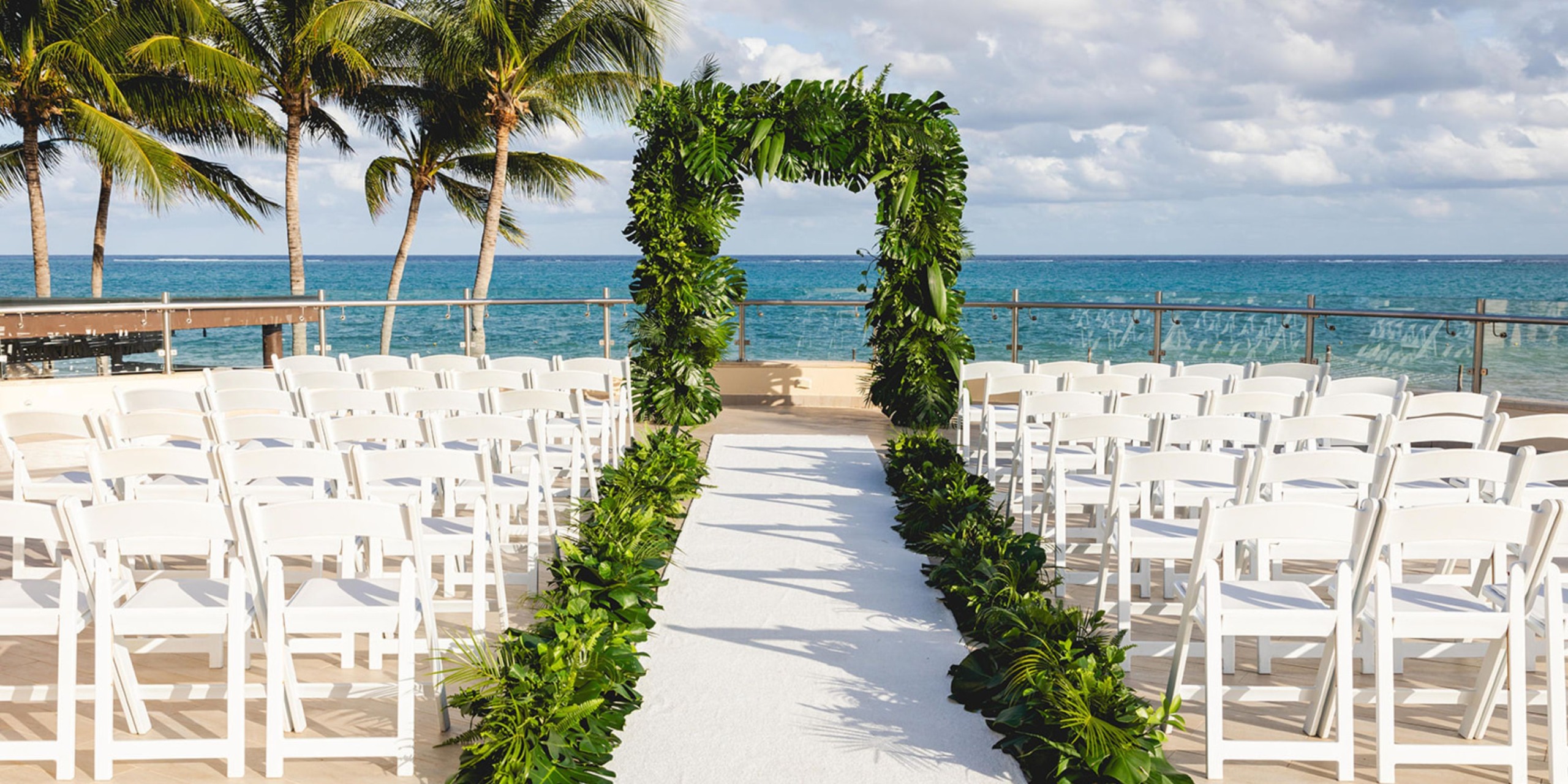 The Dreams Riviera Cancun offers a stunning destination wedding setup, featuring elegant white chairs and a lush green floral arch.
