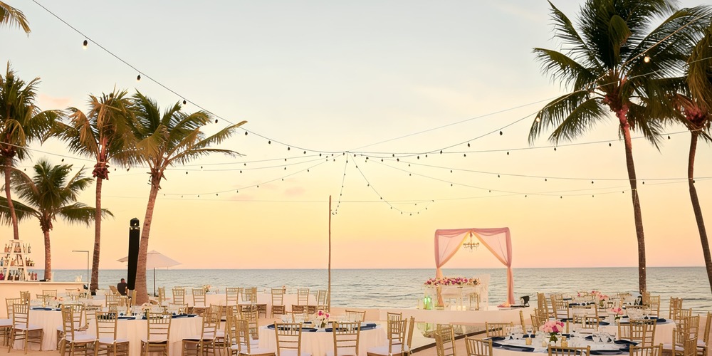 Dreams Tulum offers a stunning destination wedding experience with a beachfront setting, complete with an altar, elegantly arranged tables, enchanting lights, and palm trees at sunset.