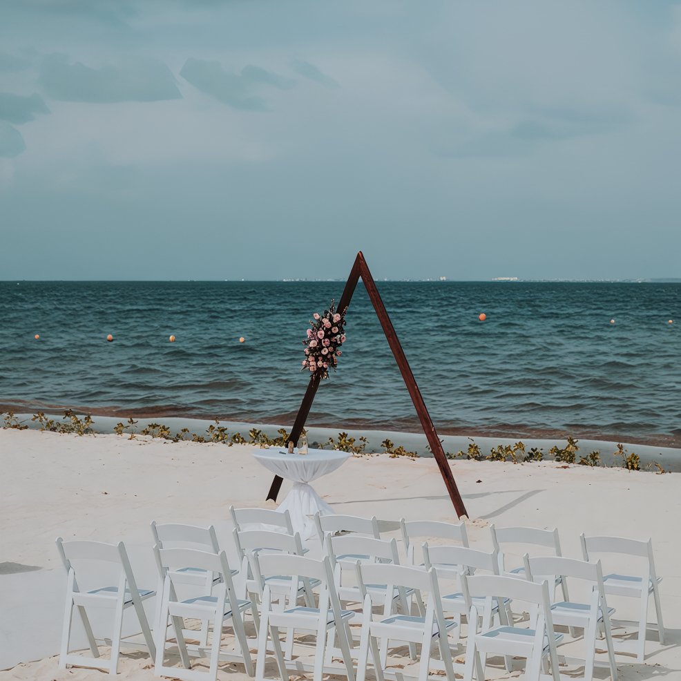 White chairs face a flower-adorned triangular arch on the sand for a destination wedding by the sea.
