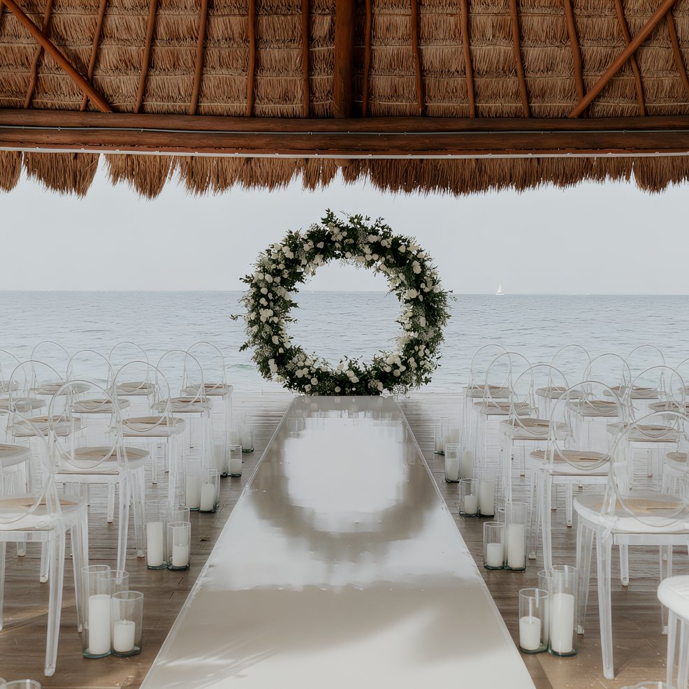 A destination wedding setup featuring clear chairs, white flowers, and candles overlooking the ocean.