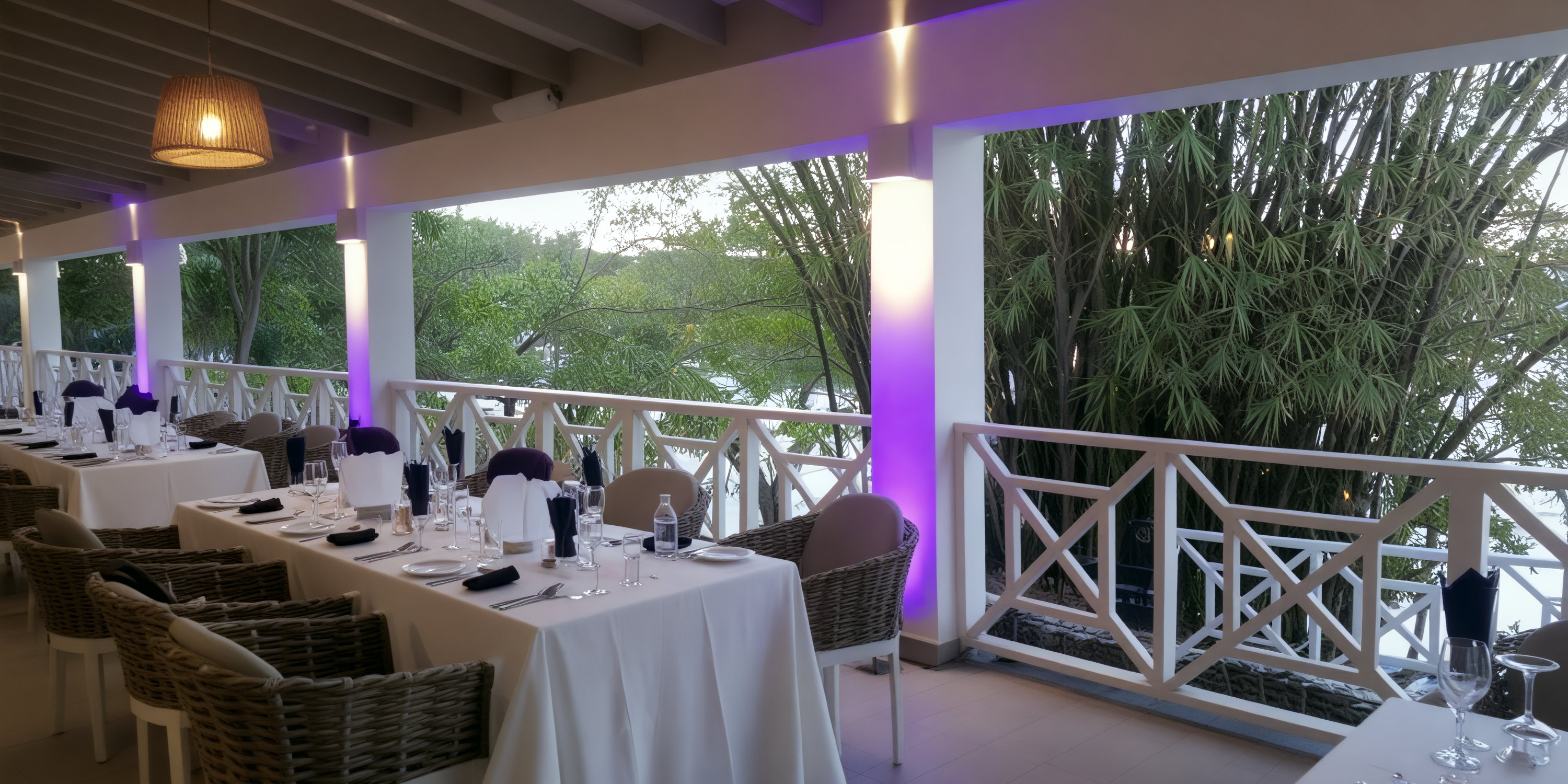 Tables and wicker chairs set for a destination wedding on Grand Palladium Jamaica’s terrace, lit in purple.