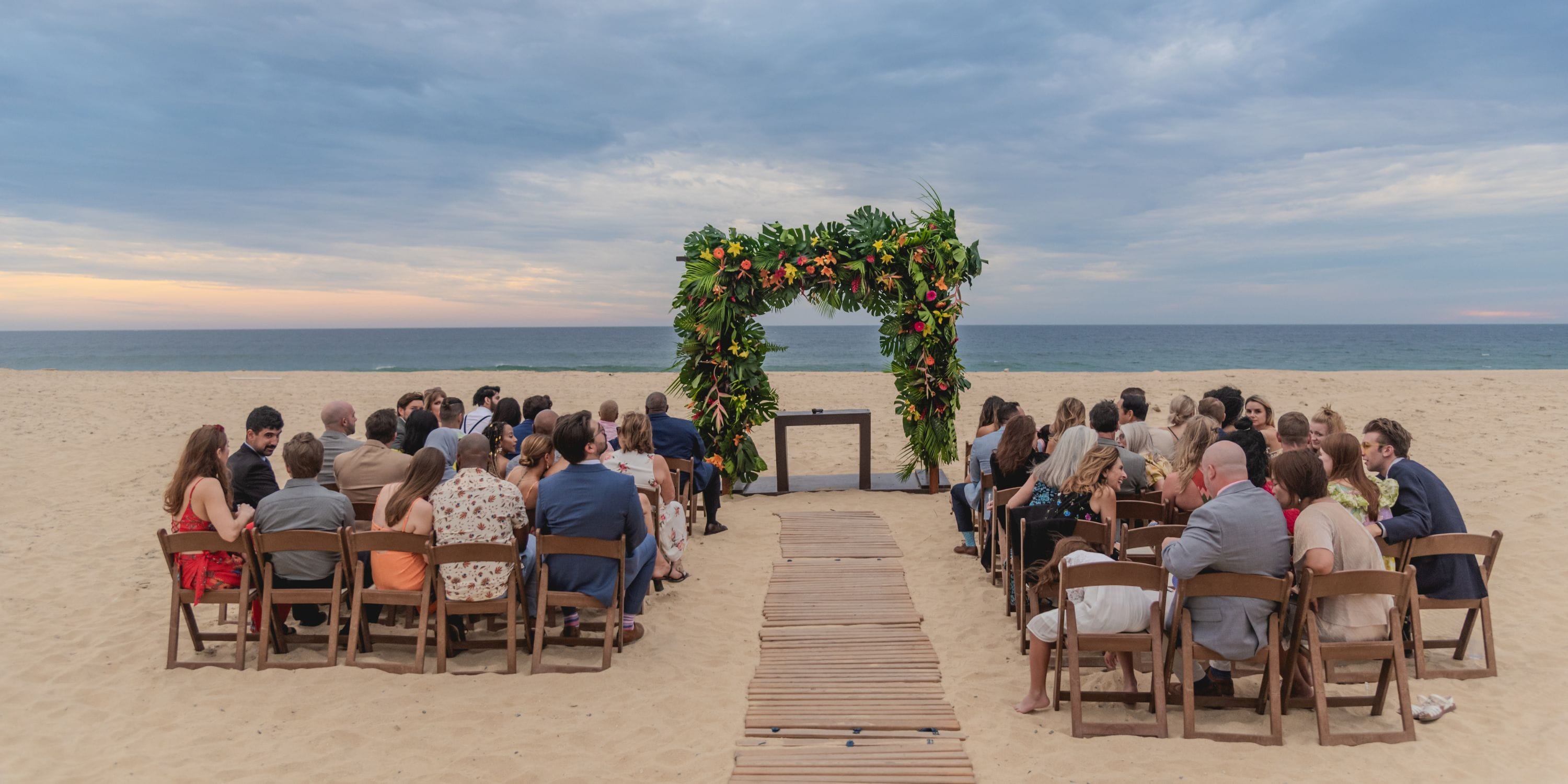 Guests at Hard Rock Los Cabos attend a destination wedding beneath a floral arch by the cloudy beach.