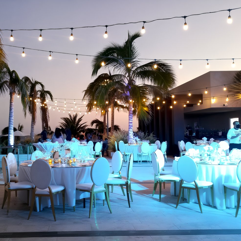 A destination wedding setup with round tables and string lights beneath palm trees at dusk.