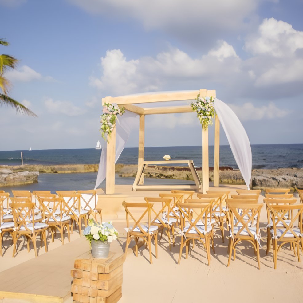 Destination wedding setup on the beach with a wooden arch, flowing white drapes, and neat wooden chairs.