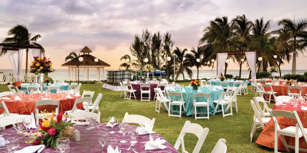At Hyatt Ziva Rose Hall, experience a breathtaking seaside destination wedding setup complete with tables, chairs, and string lights beautifully illuminated against the sunset backdrop.