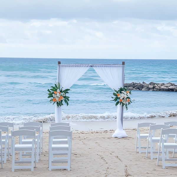 White chairs and a floral arch arranged for a destination wedding on the sandy beach by the ocean.