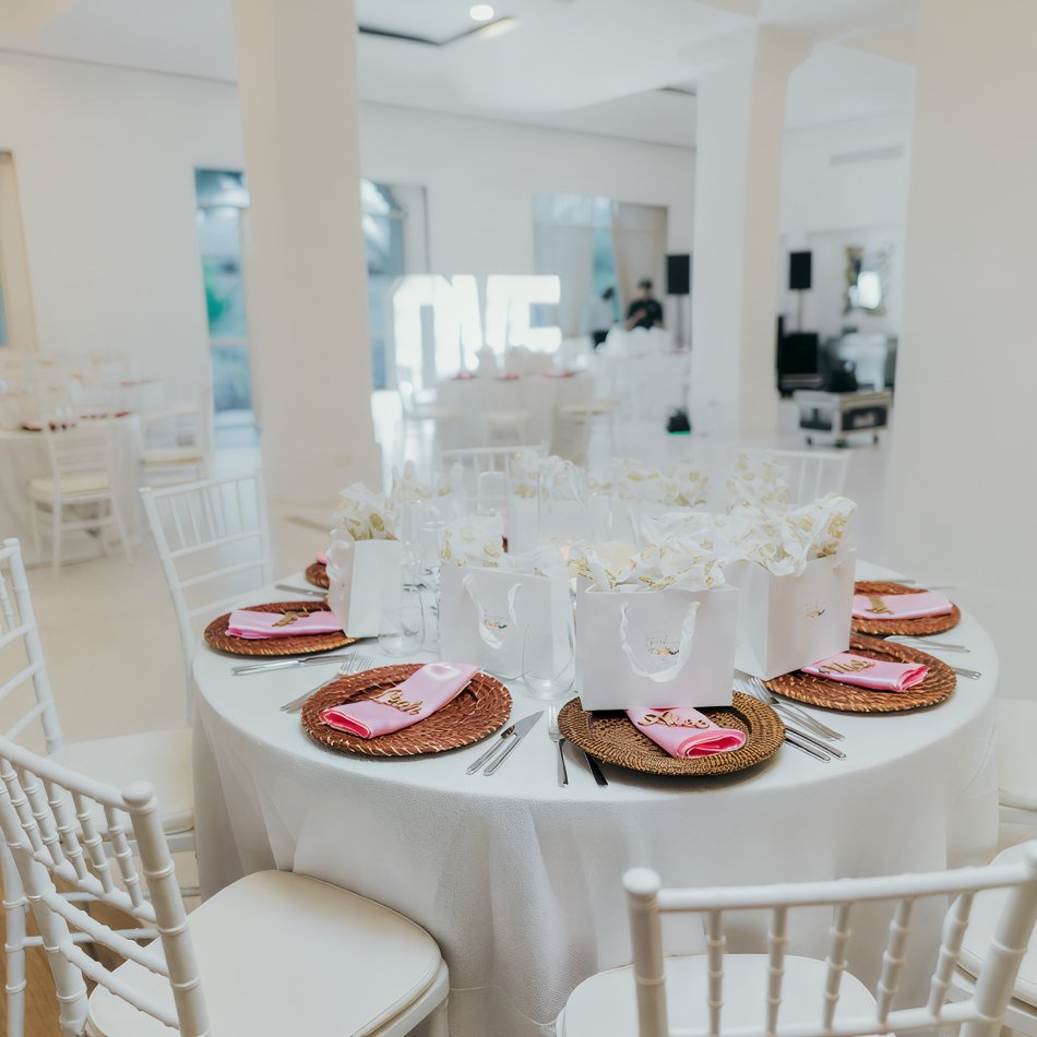 Elegant round table set for a destination wedding with white chairs, gift bags, and pink napkins in a bright room.