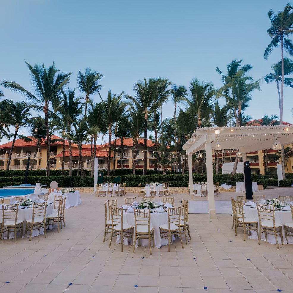 Destination wedding setup featuring round tables and chairs by a pool, surrounded by palm trees.