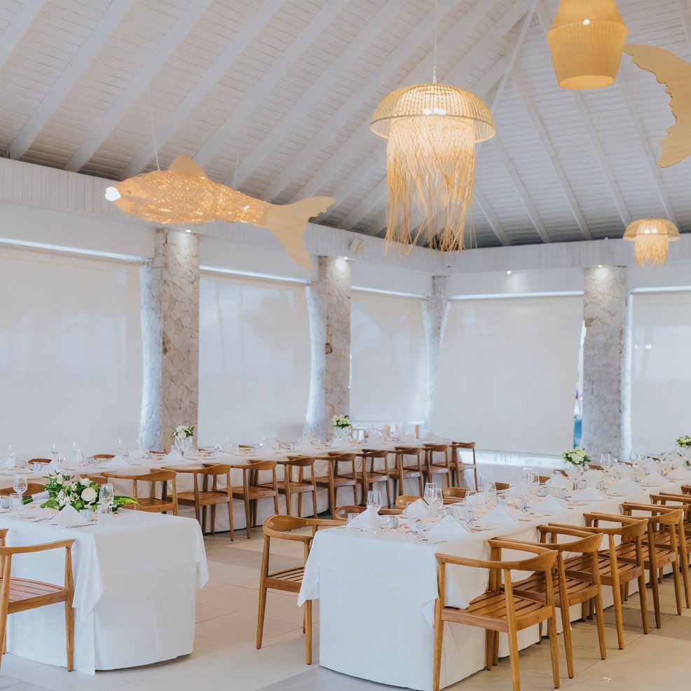 Long tables with white linens and wooden chairs set for a destination wedding in a bright, decorated room.
