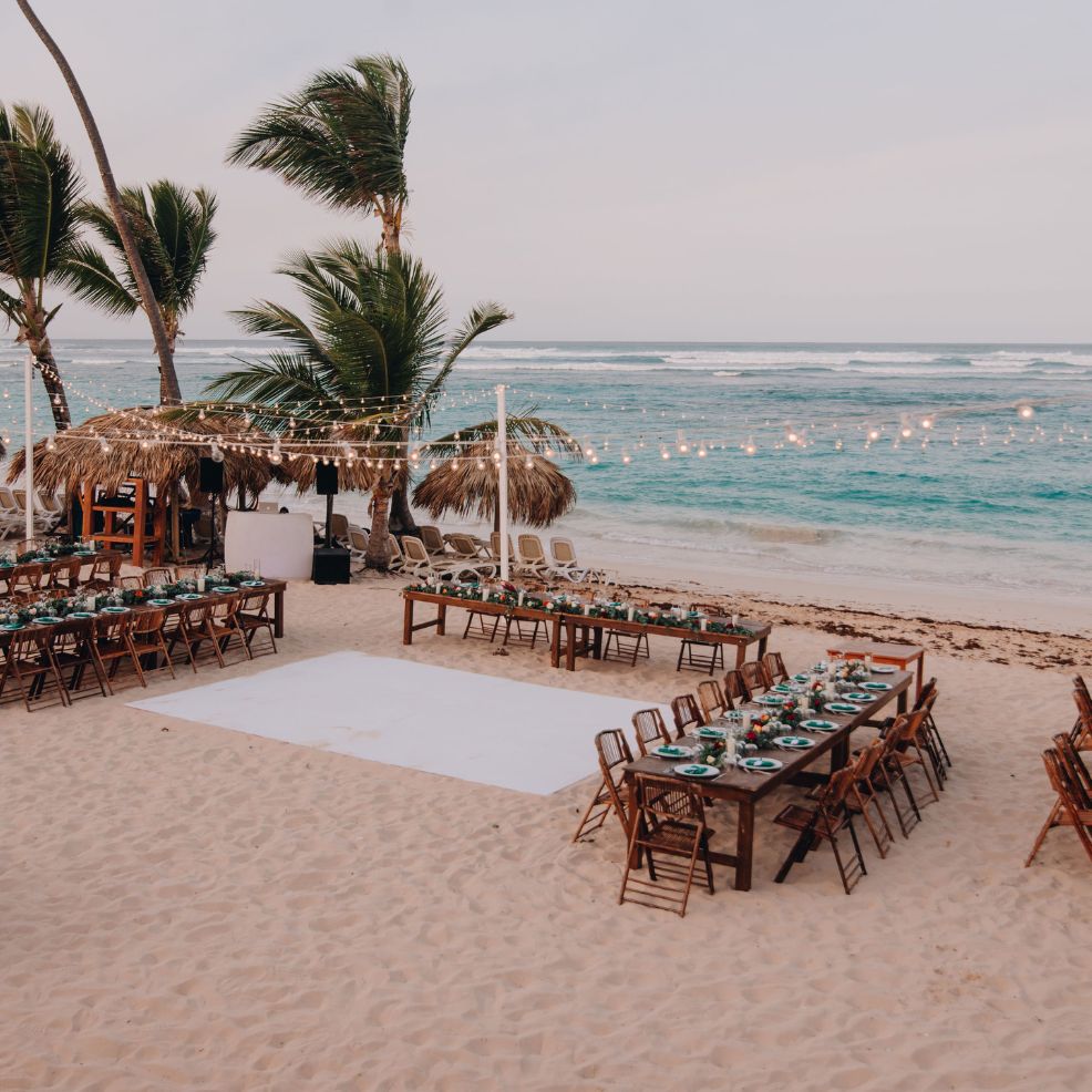 Destination wedding reception outdoors with long tables, string lights, and the ocean visible behind.