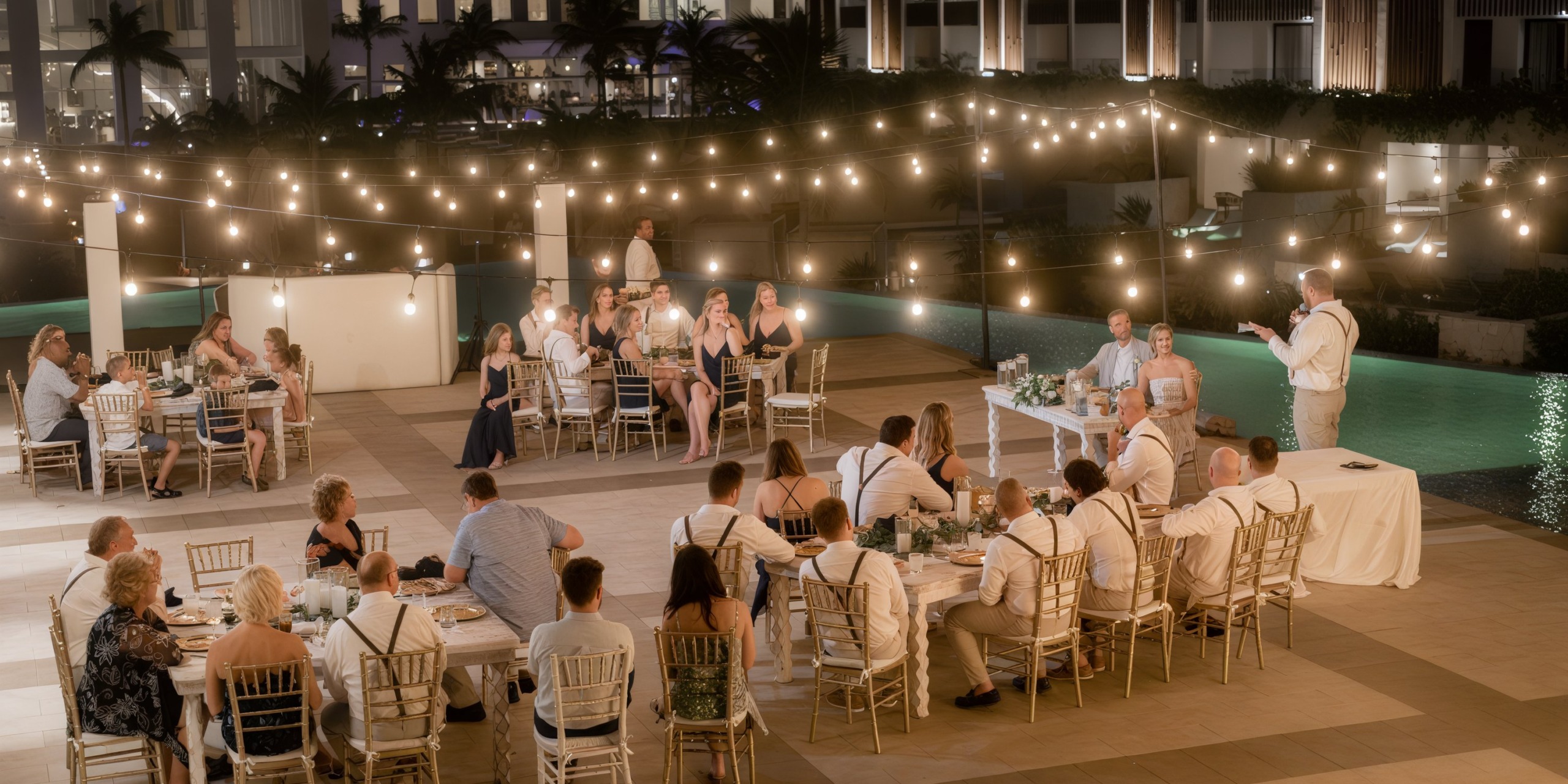A destination wedding at Majestic Elegance Costa Mujeres features an enchanting evening celebration with guests gathered by the pool, all aglow under a canopy of string lights.