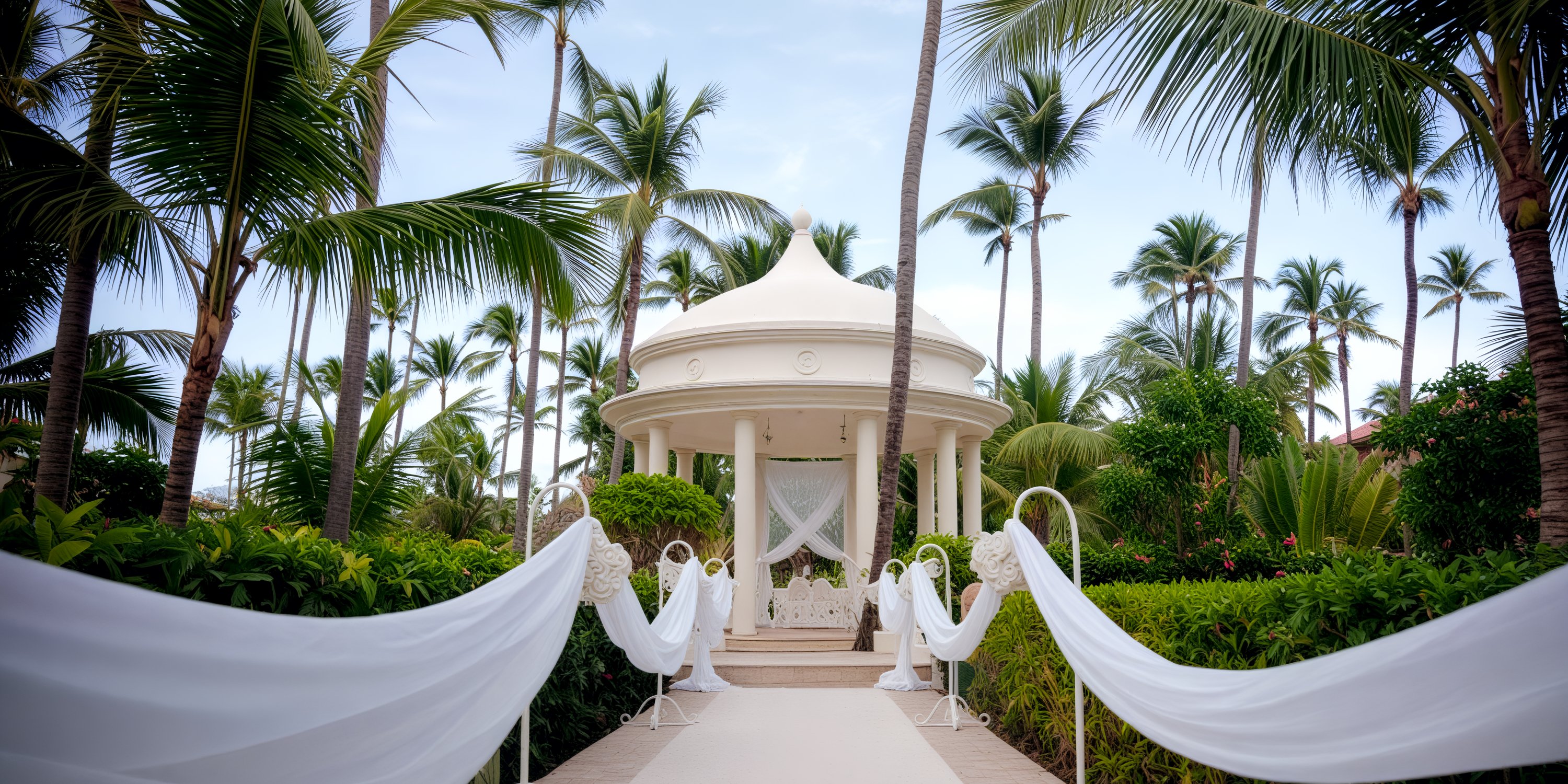 Elegant white gazebo with draped fabric among palm trees, perfect for a destination wedding setting.