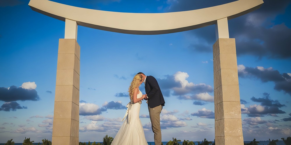 A couple kissing under the large stone archway of Majestic Elegance Costa Mujeres, set against a blue sky with clouds.