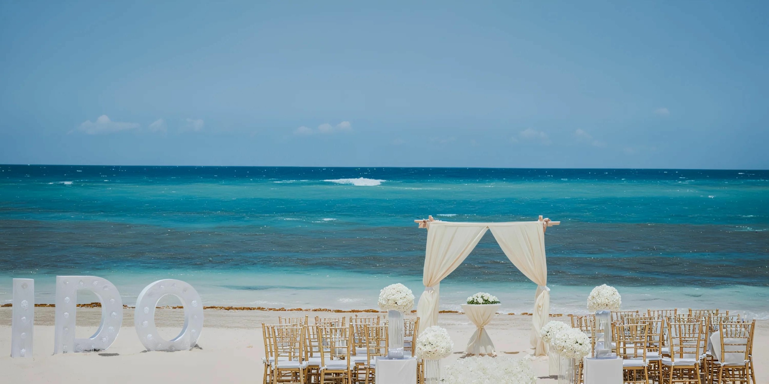 An outdoor beach wedding setup with a white canopy, chairs, and floral decorations facing the ocean. Large white letters spelling "I DO" are displayed on the left side.