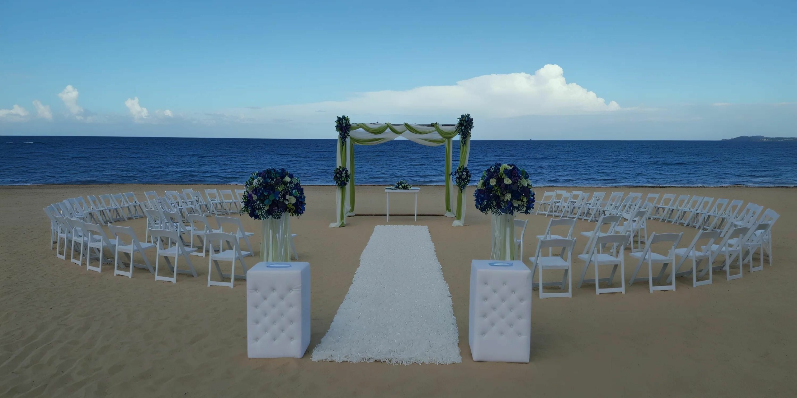 A beach wedding setup with white chairs arranged in rows facing an altar decorated with green and white fabric, and two floral arrangements on either side of the aisle runner.