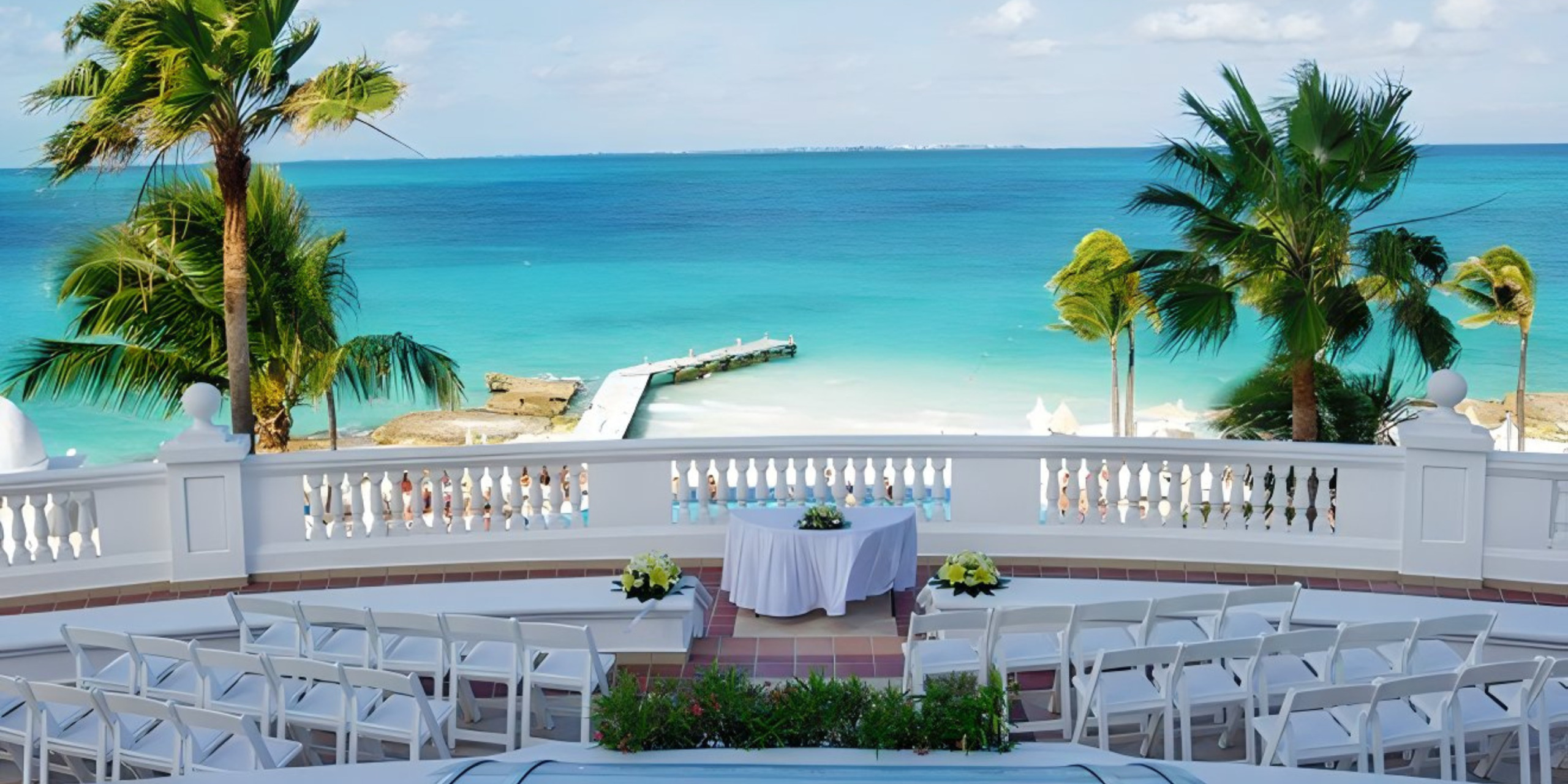 View of a destination beach wedding setup with white chairs and a table, overlooking a turquoise sea, palm trees, and a pier, from a terrace.