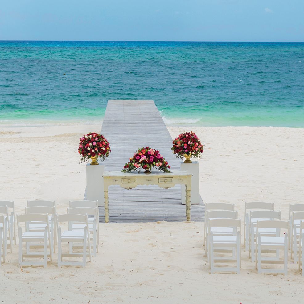 White chairs overlook a floral-adorned table and aisle on the sandy beach for a destination wedding.
