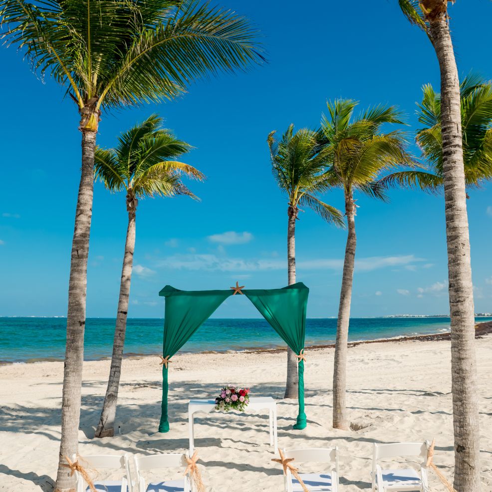 A destination wedding arch and chairs arranged on a sandy beach with palm trees and a blue sky.