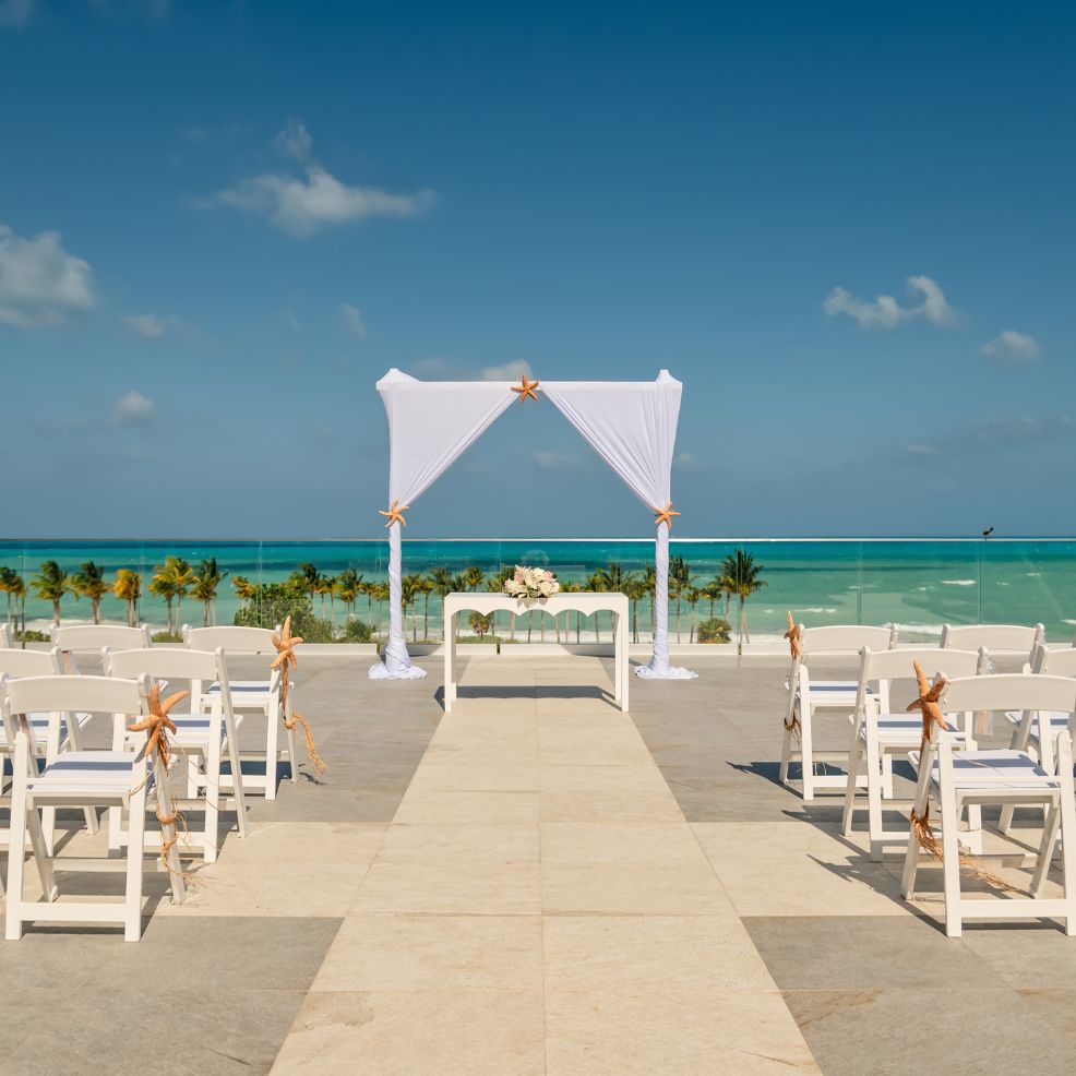 Destination wedding ceremony arrangement with white chairs and arch overlooking the ocean on a sunny day.