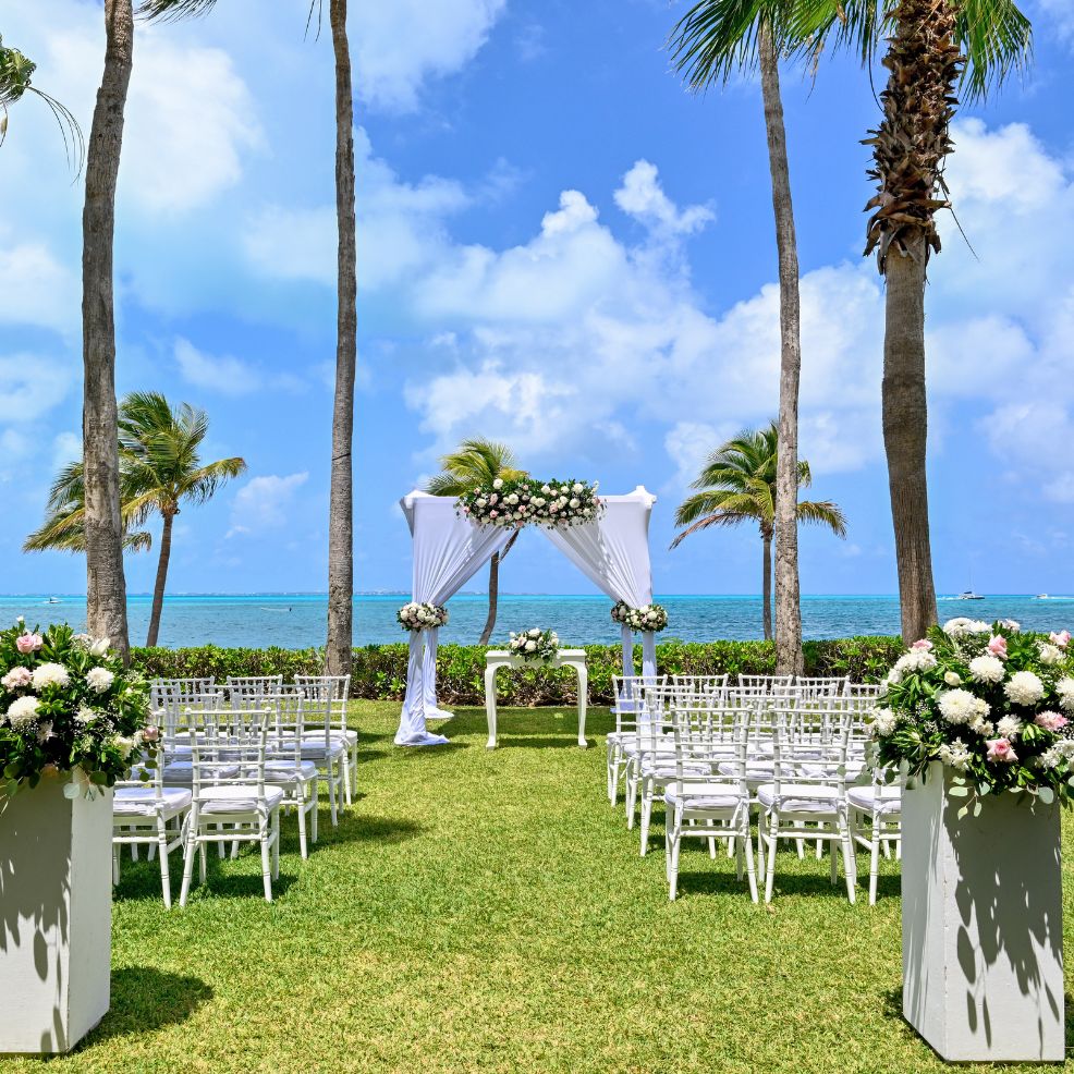 Destination wedding arrangement featuring white chairs and a floral arch by the ocean beneath palm trees.