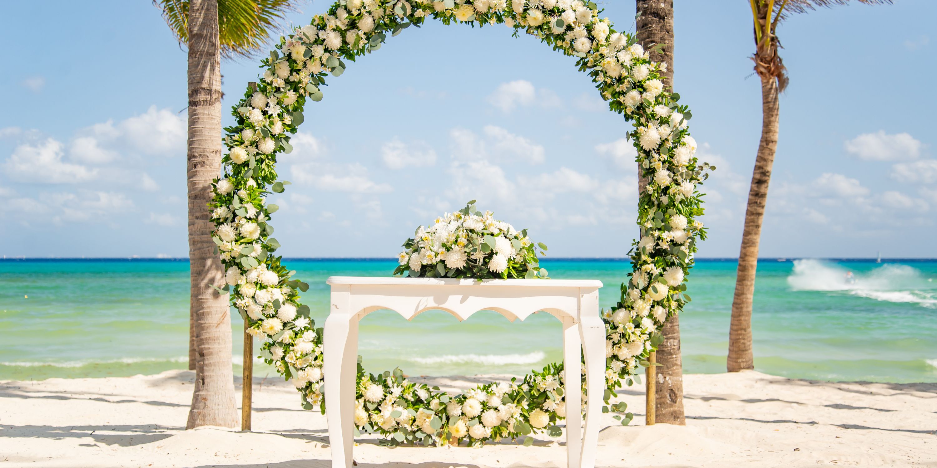 A wedding arch decorated with white flowers and a table on a sandy beach at a destination wedding.