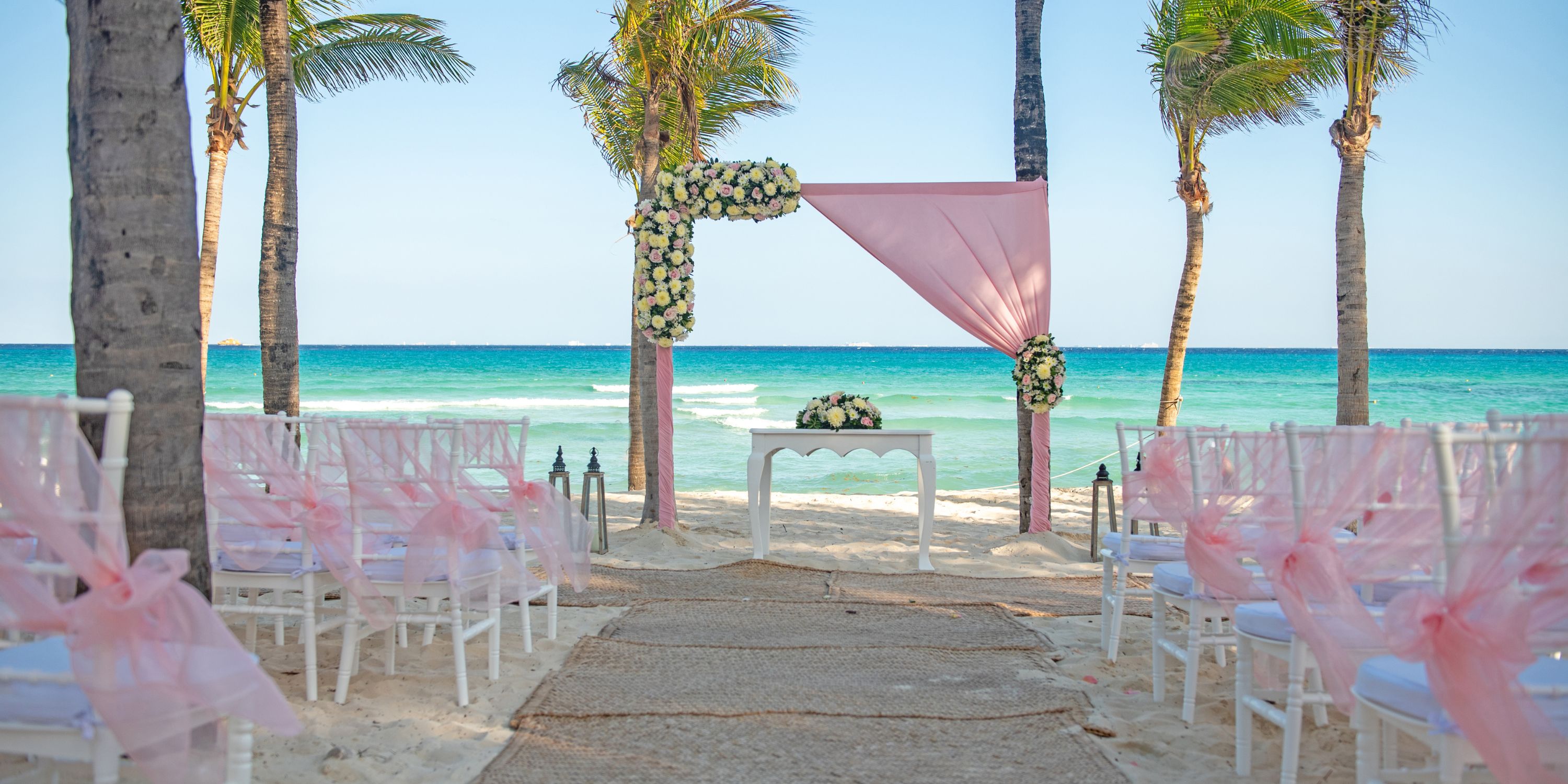 Destination wedding setup on the beach with white chairs, pink accents, and a floral arch overlooking the ocean.
