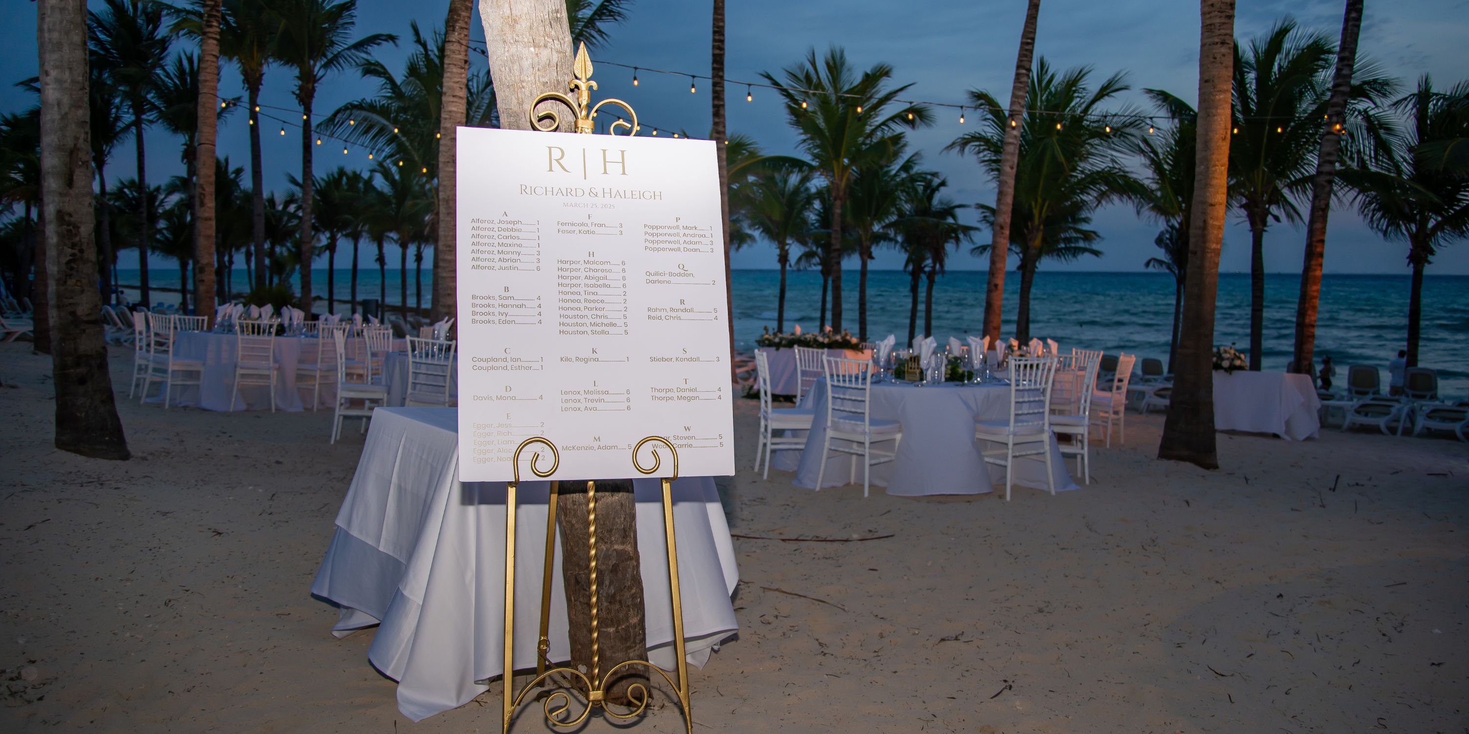 Destination wedding seating chart displayed on an easel at sunset, surrounded by tables and palm trees.