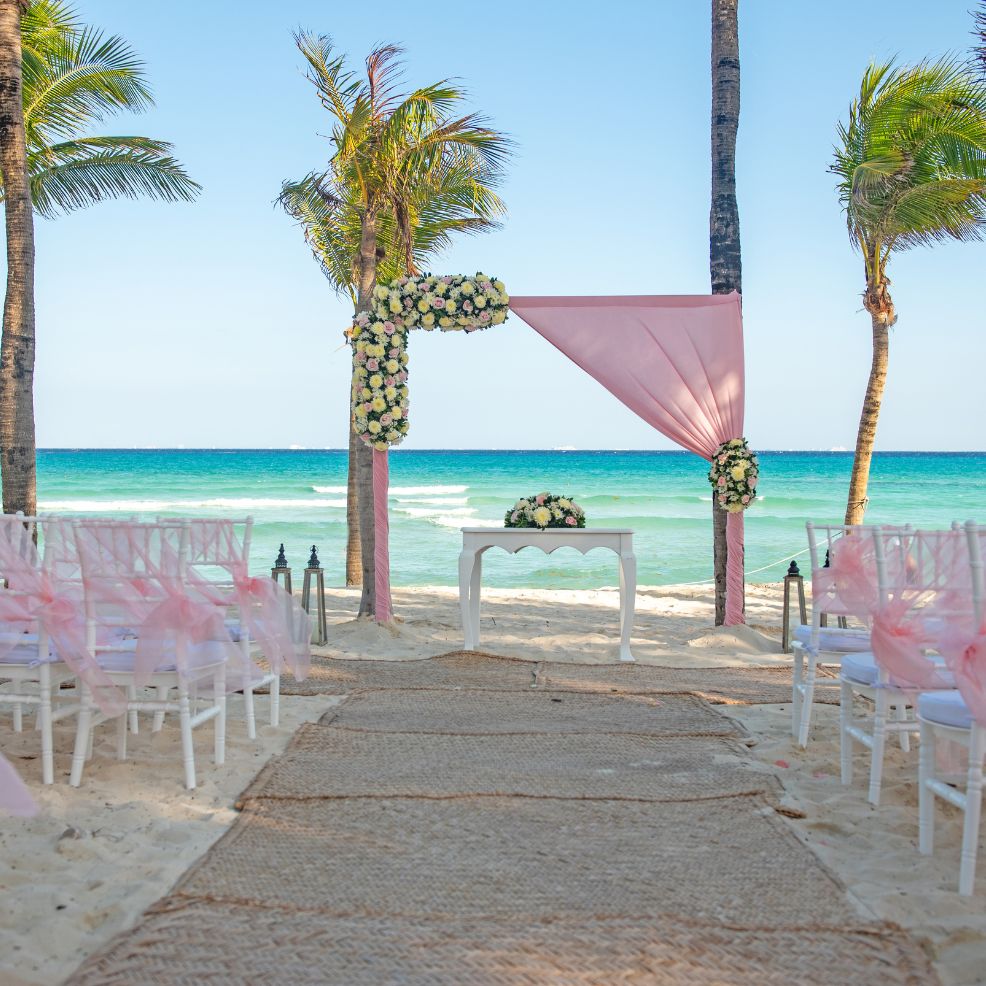 Destination wedding setup on the beach with pink and white decor, overlooking the ocean between palm trees.