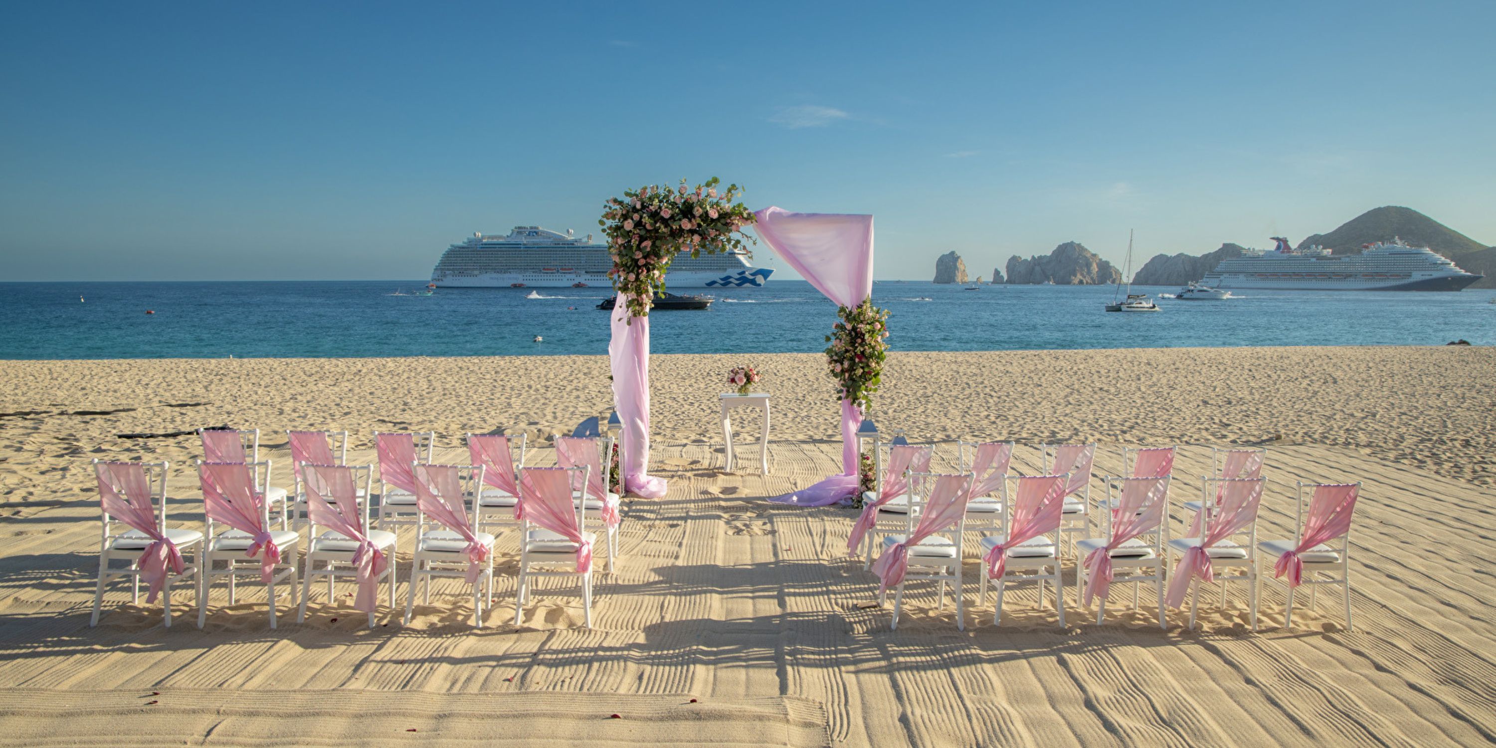 A destination wedding altar with pink chairs on the sandy beach at Hotel Riu Santa Fe, ships behind.
