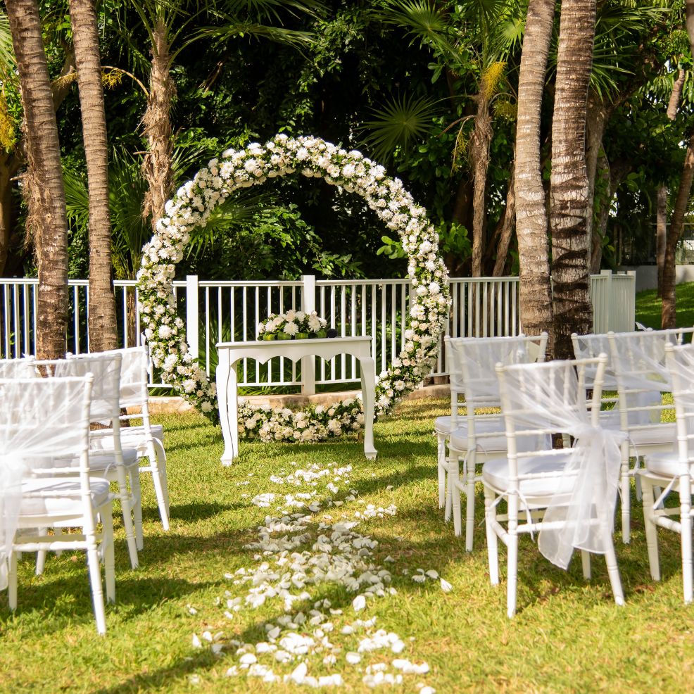 Destination wedding ceremony featuring white chairs, a floral arch, and flower petals scattered on grass.