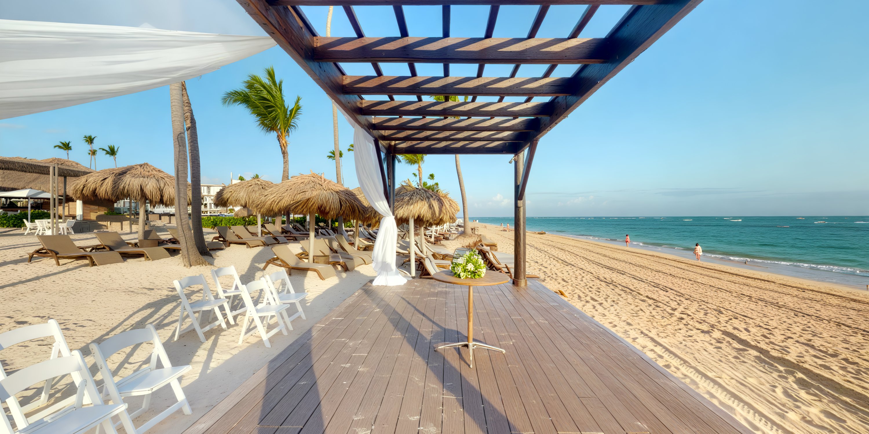 Wooden pergola with white drapes on a sandy beach, perfect for a destination wedding setup; chairs and palm trees in the background.