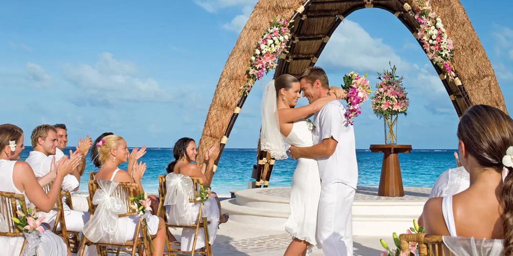 A bride and groom kissing in front of a Dreams Riviera Cancun on the beach.