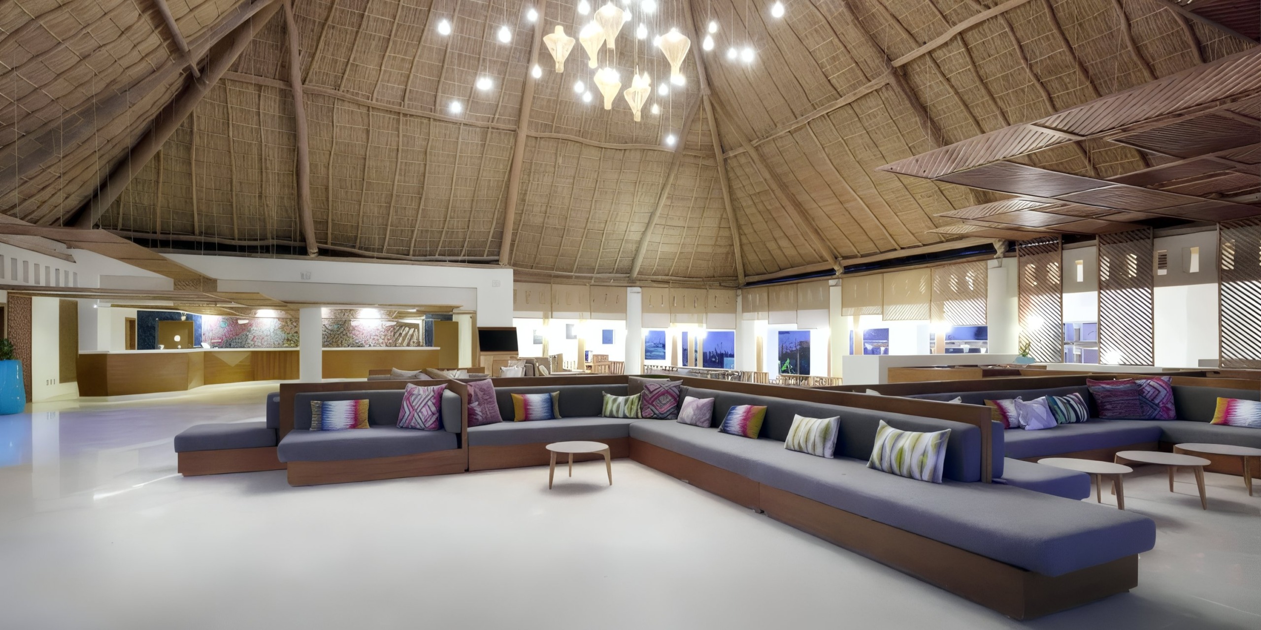 The Grand Palladium Vallarta offers a spacious lobby featuring a large L-shaped sectional sofa adorned with colorful pillows, along with a high thatched ceiling and a modern chandelier.