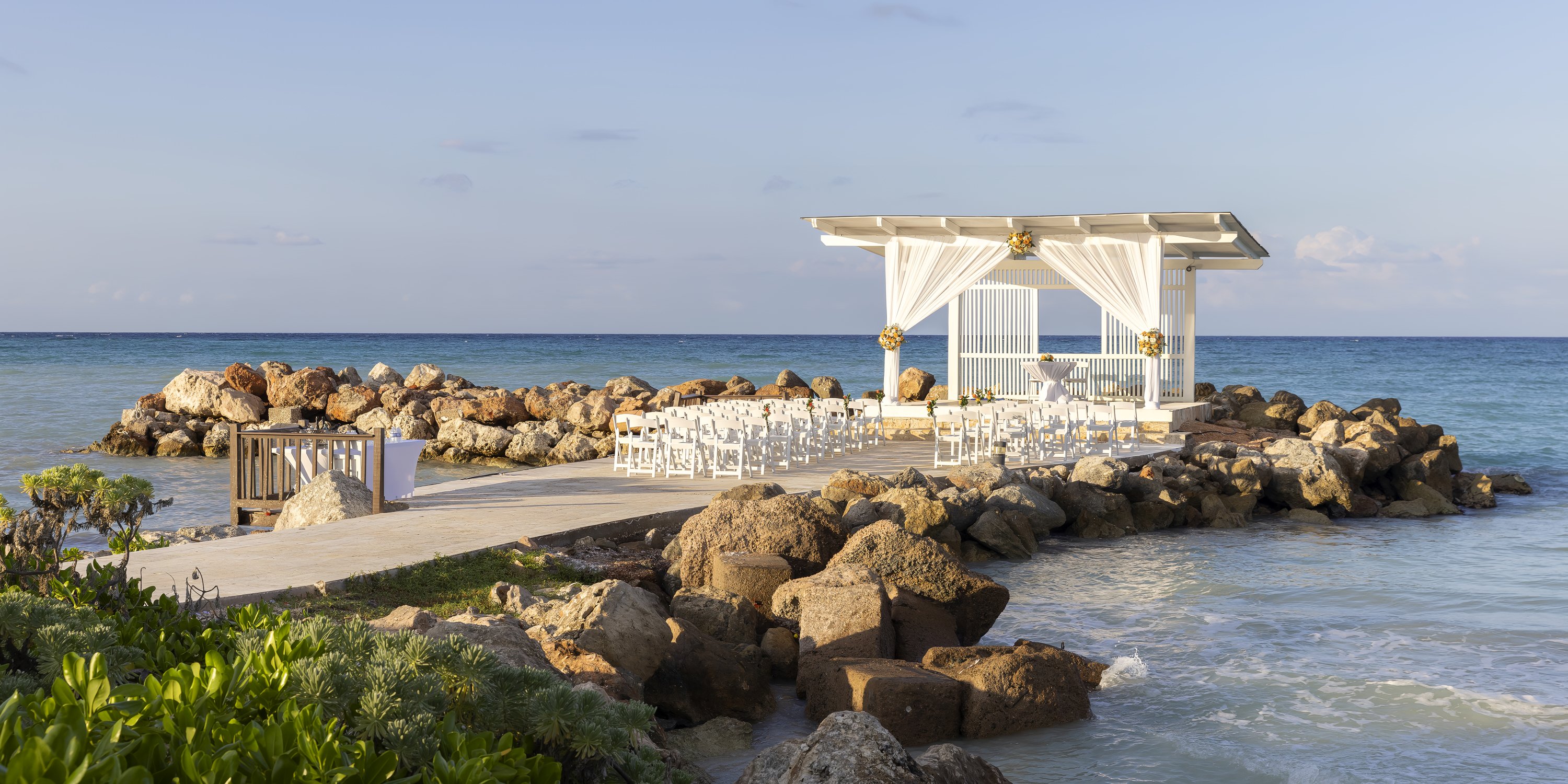 Destination wedding setup at Royalton Blue Waters on a rocky pier by the ocean under a clear sky.