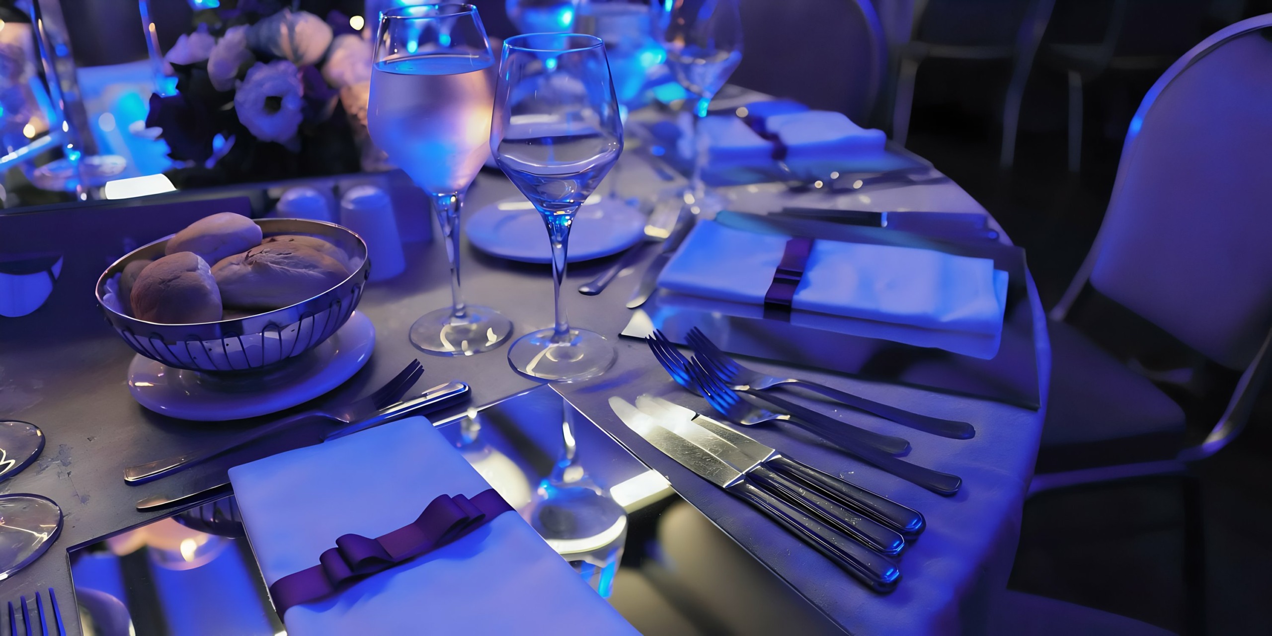 A formal dining table setting with silverware, glassware, and a bread basket under blue ambient lighting at Majestic Elegance Punta Cana.