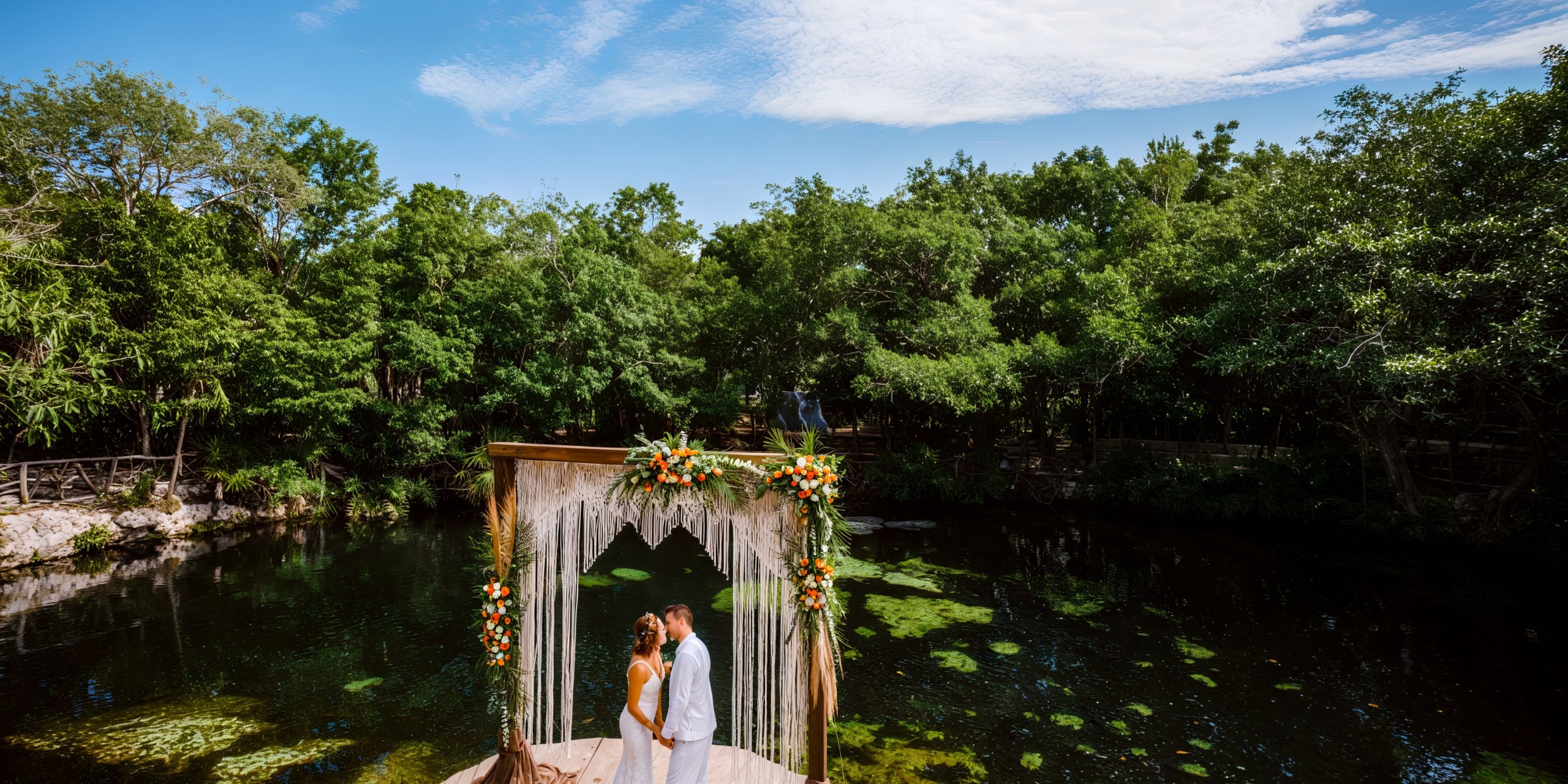 A couple stands beneath an arch at the lush pond of Sandos Caracol Eco Resort, celebrating their destination wedding.