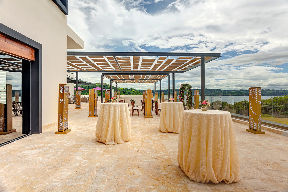 An outdoor event space at one of the finest Costa Rica wedding venues, featuring tall cocktail tables covered in beige tablecloths. The area has a partial covering and overlooks a scenic view of green landscape and water under a partly cloudy sky.