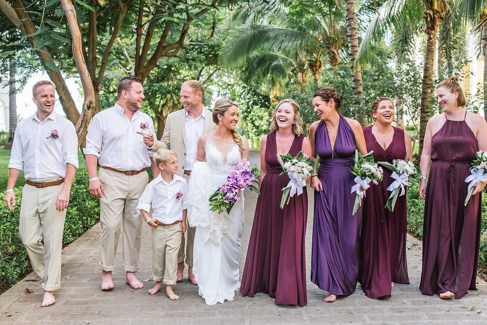 A bride and groom walk with their bridal party, including a young boy and five adults in purple dresses, outdoors among the lush greenery typical of Costa Rica wedding venues. They are all smiling and appear relaxed and joyful.