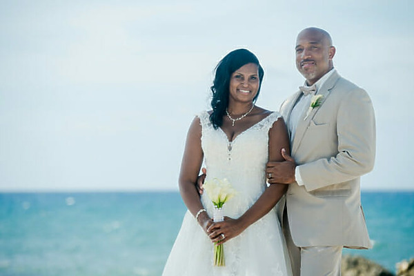 A bride in a white dress and a groom in a beige suit stand together, smiling, in front of the ocean during their wedding in Jamaica. The bride holds a bouquet of white flowers.