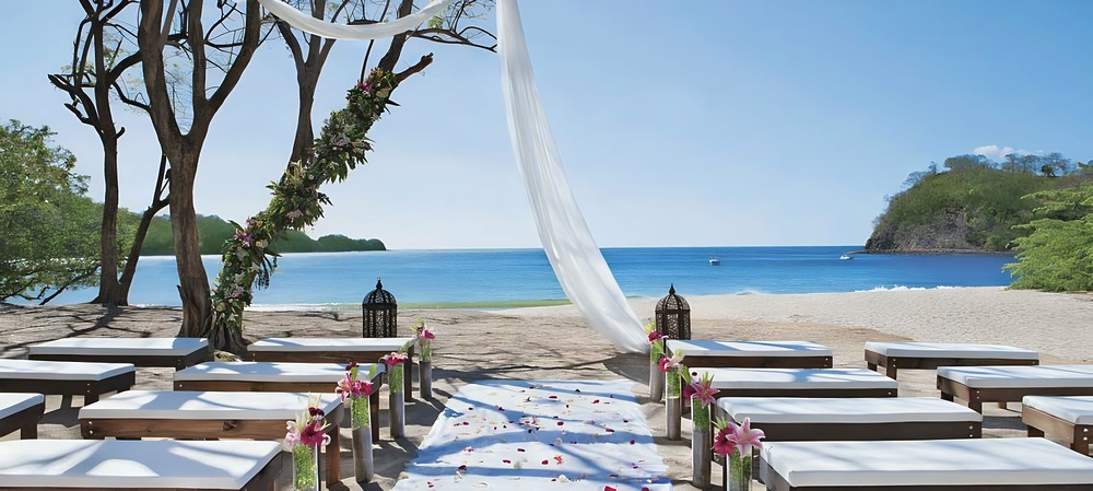 A beach wedding setup with white benches, floral decorations, and a fabric-draped arch facing the ocean on a sunny day is just one of the stunning Costa Rica wedding venues.