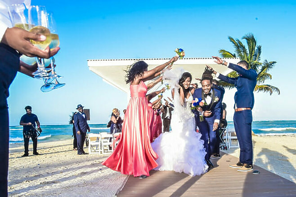 A newlywed couple walks down the aisle on a beach in Jamaica as guests cheer and hold up flower bouquets. The bride wears a white dress, and the groom is in a dark suit. Palm trees and the ocean are in the background.