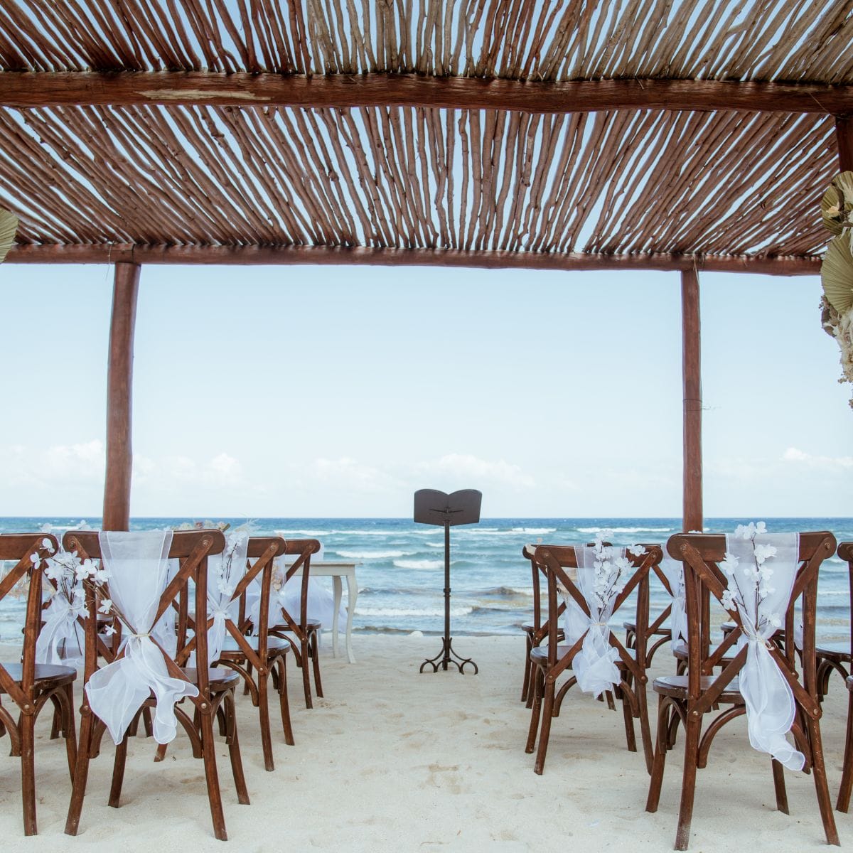 Wooden chairs with white accents are arranged for a destination wedding under a beachside pergola.