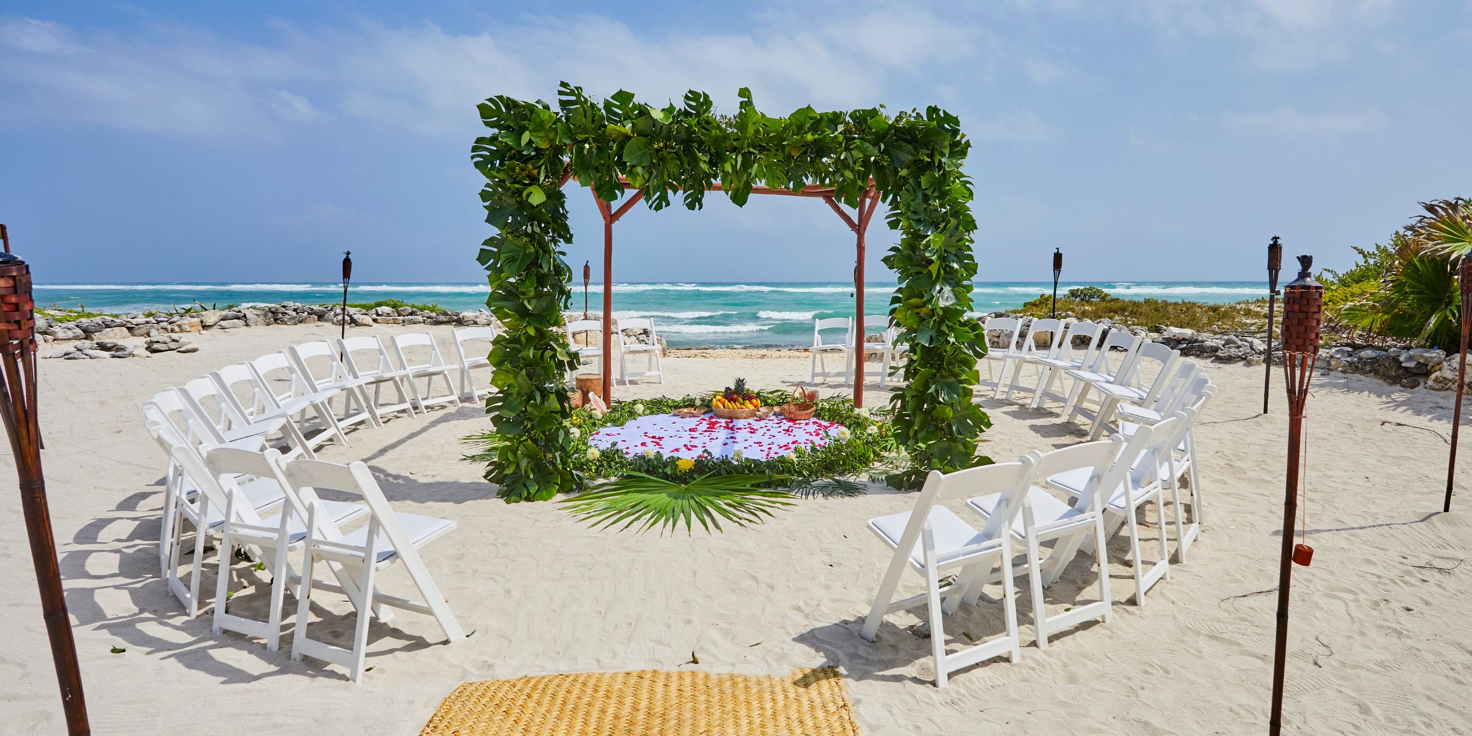 Destination wedding setup with a circular arrangement of white chairs and leafy arch at Bahia Principe Explore Coba beach.
