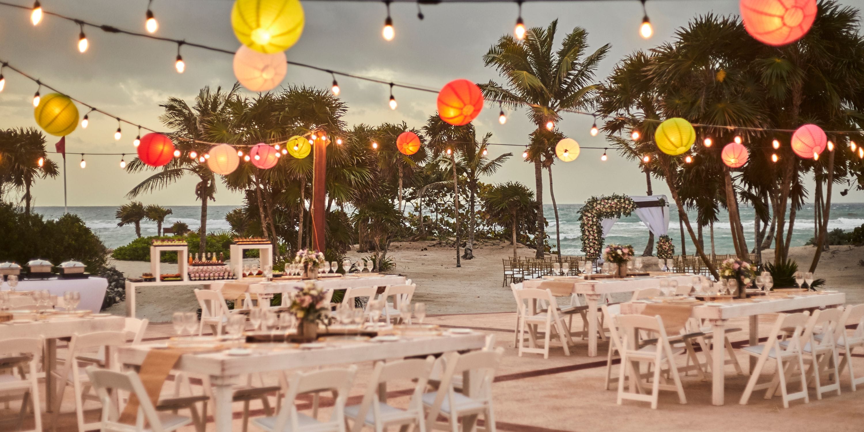 Destination wedding setup on the beach at sunset, with decorated tables, string lights, and palm trees.