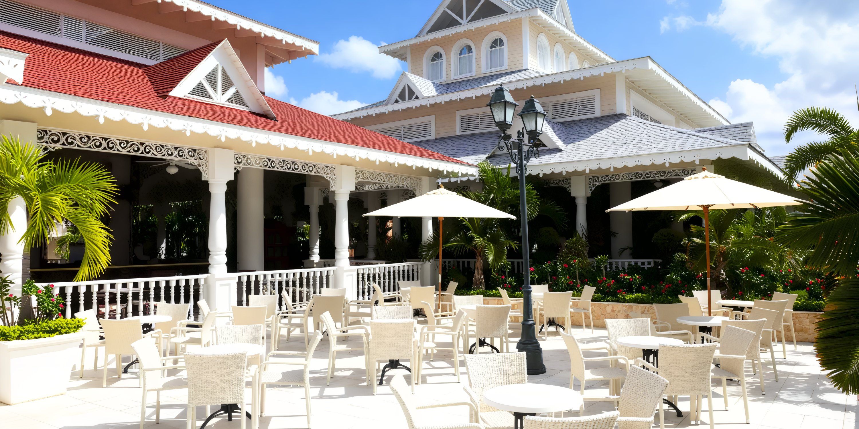 The patio at Bahia Principe Luxury Bouganville, adorned with white tables and chairs amid lush tropical plants and ornate buildings, offers a picturesque setting perfect for a destination wedding.