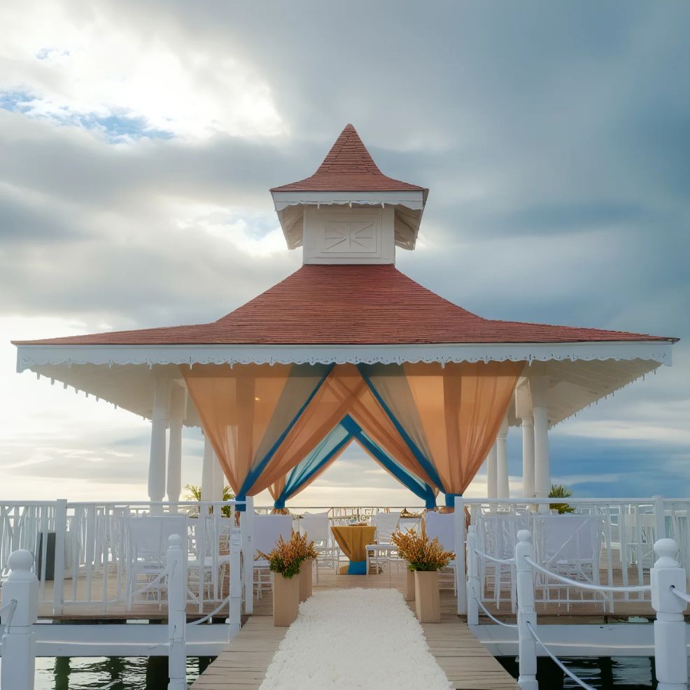 A white gazebo with a red roof on a boardwalk, decorated with orange and blue drapes, set up for an event. White chairs and potted plants are placed along the aisle.
